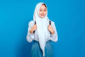 A Cheerful Asian girl wearing school uniform and hijab, smiling confidently while holding backpack straps. Positive and friendly facial expression on blue background photo
