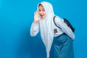 A Cheerful Muslim schoolgirl in uniform and hijab making a listening gesture with playful smile and expressive face. Back to school theme on isolated blue background. photo