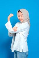 A Confident young Asian woman wearing hijab smiling proudly while showing muscle flexing gesture. Happy face expression symbolizing strength and empowerment. photo