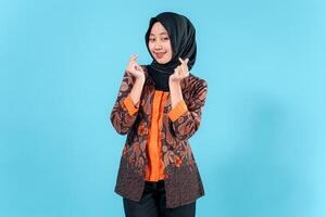 A Smiling woman wearing a hijab and traditional batik outfit makes a finger heart gesture, expressing joy and friendliness in a studio portrait. photo