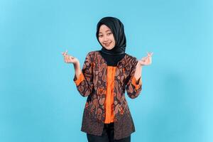 Smiling young Asian woman wearing a hijab and traditional batik outfit makes a finger heart gesture, expressing joy and friendliness in a studio portrait. photo