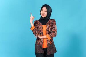 Confident young Asian woman wearing hijab and batik outfit smiling brightly while pointing upward with one finger, expressing excitement, inspiration, or having a great idea. photo