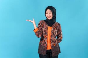 A Young Asian Muslim woman wearing a batik outfit and hijab smiling confidently while pointing sideways with open palm, showing a cheerful and welcoming facial expression. photo