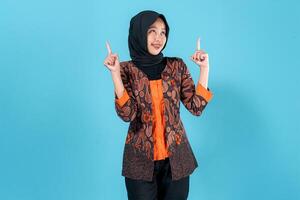 A smiling young Asian woman wearing a hijab and traditional batik outfit points both index fingers upward, showing a cheerful and enthusiastic facial expression in a studio portrait. photo