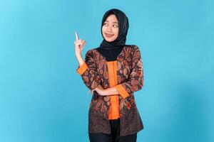 A Confident young Asian woman wearing hijab and batik outfit smiling brightly while pointing upward with one finger, expressing excitement, inspiration, or having a great idea. photo