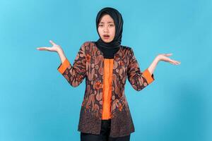 Asian woman wearing a hijab and batik shirt looks confused and surprised with both hands raised, expressing uncertainty or doubt against a solid blue background. photo