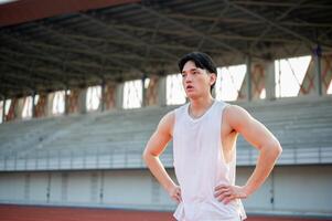 Asian man in white tank top is standing with hands on waist taking a rest after running on racetrack photo