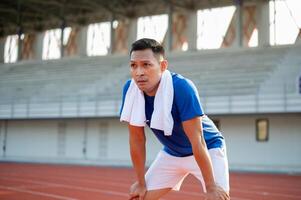 Asian man in blue t shirt standing with hands on his knee, taking a rest after running on racetrack. photo