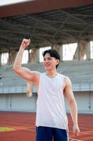 An asian man in white tank top is smiling and raising a fist while standing on the racetrack. photo