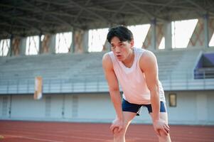 Asian man in white tank top standing with hands on his knee, taking a rest after running on racetrack. photo