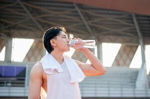 Asian man in white tank top standing and drinking from a water bottle, taking a rest after running. photo