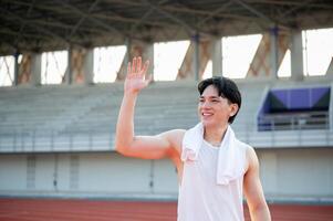 An asian man in white tank top is smiling and waving a hand while standing on the racetrack. photo