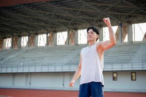 Asian man in white tank top is closing eyes and raising a fist while standing on racetrack. photo