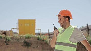 Construction Supervisor in Safety Vest and Helmet Uses Walkie-Talkie While Pointing at Worksite Area video