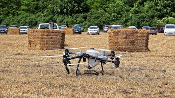 Agricultural drone standing on dry field near hay bales and parked cars at farming event, showcasing drone technology used in precision agriculture and smart video