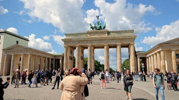 Berlin Germany 25.05.2025, Crowds of tourists gather around Brandenburg Gate under blue sky with clouds, iconic Berlin landmark with people walking, taking video