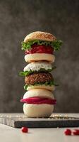 A stack of four colorful burgers on a wooden board. The burgers include lettuce, tomato, and a variety of patties. The background is neutral and textured. photo