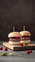 Two mini burgers on a wooden board. Each burger has a sesame seed bun, a beef patty, and a slice of tomato. Fresh cranberries and green leaves are scattered around. photo