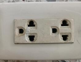 Close up of double electrical outlet with worn and dirty appearance, mounted on white surface, showing signs of age and use, with some dust and minor damage visible photo