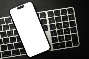 A modern smartphone with a blank white screen rests on a white and black computer keyboard, ideal for showcasing digital content or technology concepts. Copy space. photo
