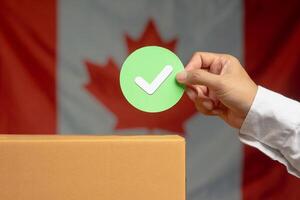 Hand holding a green check mark symbol overhead the voting box at place election with the Canada flag background photo