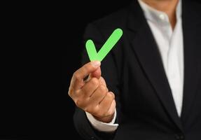 Close-up of a businessman holding a green check mark symbol while standing on a black background photo