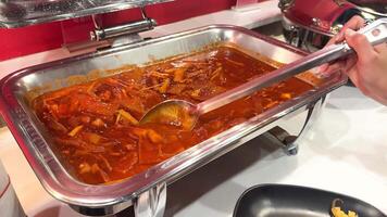 A person serves spicy Korean tteokbokki from a buffet tray onto a plate in a well-lit setting video