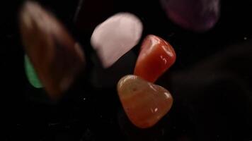 Vibrant chakra stones tumble onto a black surface, showcasing their colors and textures in slow motion under soft lighting video