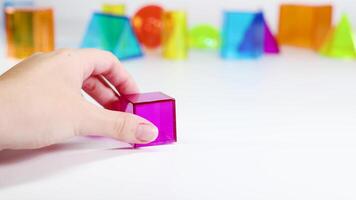 A hand manipulates a pink cube among vibrant 3D shapes on a white surface, highlighting geometry and color video