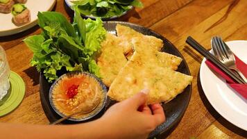 Hands reach for crispy shrimp pancakes with greens and sauce on a wooden table. Bright lighting enhances the vibrant colors video