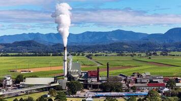 Aerial footage of a sugarcane factory emitting smoke, surrounded by lush fields and mountains under clear skies video