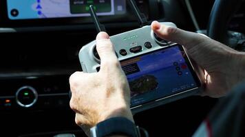 Hands operate a drone controller inside a vehicle, displaying a scenic view on the screen. Bright lighting enhances focus video