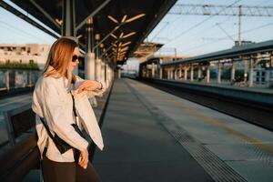 Stylish woman checking time on her watch while waiting for train at sunset, standing on platform of railway station with train arriving in background. Woman glancing at watch, waiting for train photo