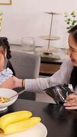 A young girl pours milk into a cereal bowl at a breakfast table, assisted by her mother in a cozy kitchen setting video