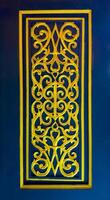 A vibrant close-up of an intricate golden carving, showcasing symmetrical, swirling traditional Thai patterns, set within a rectangular frame against a rich, deep blue background. The golden relief photo