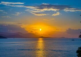A breathtaking wide-angle shot captures the sun as it dips below the horizon, casting a vibrant golden glow across the tranquil ocean. The distant mountains are silhouetted against the fiery sky photo