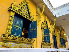 The intricate golden and blue windows of a Thai Buddhist temple facade are captured from a low-angle perspective, highlighting the ornate traditional architecture and vibrant cultural heritage. The photo