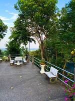 This eye-level, wide-angle shot captures a peaceful outdoor seating area with concrete benches and a table, nestled on a paved ground. Lush green trees frame the scene, offering a shaded retreat with photo