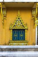 This eye-level, wide-angle shot captures the intricate details of a vibrant golden and blue window, set against a textured ochre wall of a Buddhist temple. The exquisite craftsmanship, typical of photo