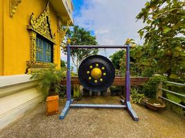 A large, black and gold traditional temple gong hangs from a rustic purple and blue wooden frame, situated on a concrete platform outside a vibrant yellow Thai temple adorned with intricate golden photo