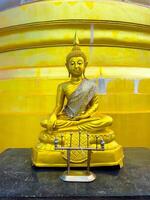 A magnificent golden Buddha statue, adorned with a shimmering silver and gold sash, sits serenely in a meditation pose atop a dark stone surface. The statue is positioned against the vibrant, luminous photo