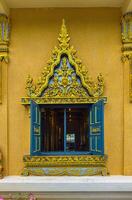 A beautifully detailed window, with its blue wooden shutters opened wide, reveals a glimpse into the richly decorated interior of a Thai temple. The window frame is exquisitely carved with intricate photo