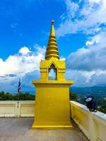 A resplendent golden Buddhist shrine, topped with a tiered spire and featuring a small mirrored window, stands prominently on a white stone terrace. Behind it, a Thai flag flutters gently, and lush photo
