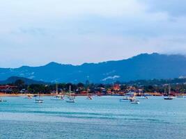 This wide-angle shot captures a serene bay filled with various boats gently resting on the tranquil, blue-green water. In the background, lush green mountains rise majestically, their peaks partially photo