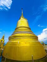 A towering, shimmering golden stupa, with its distinct tiered base and sharp spire, dominates the frame, captured from a low-angle perspective looking upwards. The brilliant gold surface reflects the photo