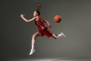 Dynamic female basketball player in mid-air jump with flying ball, wearing red uniform on gray studio background symbolizing strength, motion, and athletic performance. photo