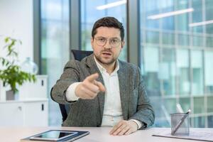 Portrait of angry young businessman holding an online meeting, talking aggressively and pointing an accusing finger at the camera. photo