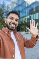 A smiling man on a call outside a modern building, waving at the camera photo