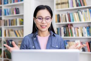 An Asian woman smiles while participating in a call, using earphones, with a library in the background. photo