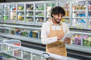 A store worker, wearing an apron, uses a tablet to manage inventory in front of refrigerated display cases, smiling as he works. photo